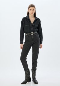 Camisa de mezclilla negra con cierre de botones, combinada con jeans negros de tiro alto. Las botas de vaquero negras presentan costuras intrincadas y puntas afiladas.