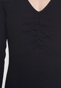 Robe noire en tissu texturé, avec un décolleté en V et un détail de fronces au centre. Manches longues et coupe ajustée.