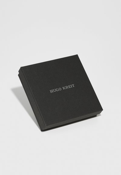 Melna cietā kastē kvadrātveida formā ar gludu tekstūru. Priekšpusē ir uzspiedis sudraba teksts: "HUGO KREIT." Minimalistisks dizains bez papildu rakstiem.