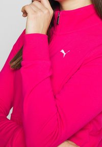 Personne portant un haut zippé Puma rose vif avec logo blanc, main reposant près du visage, recadré au niveau du cou et du milieu du torse.