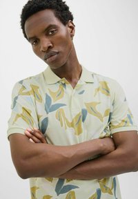 Camisa polo de manga corta, de color verde claro, con un patrón abstracto en tonos azules y amarillos. Presenta un cuello clásico y una textura de tejido suave.