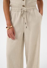 Femme portant un pantalon large en lin beige avec taille à cordon et poches, associé à un haut boutonné assorti et un bracelet doré au poignet gauche.