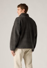 Veste polaire gris foncé avec col montant, surface texturée et coupe décontractée, associée à un pantalon de couleur claire.