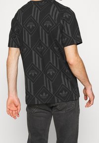 Hombre con una camisa negra de manga corta con patrones geométricos oscuros y logotipos, combinado con jeans gris oscuro, visto desde la espalda.