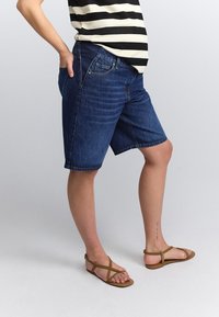 Mørkeblå denim Bermuda-shorts med almindelig pasform, der har fem lommer og opsmøgede kanter. Båret med tanfarvede remsandaler.