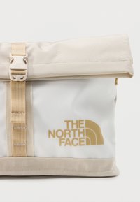 Μπεζ και λευκό σακίδιο με ρολό κορυφή, με μπεζ λουρί πόρπης και το λογότυπο "THE NORTH FACE" σε ανοιχτό καφέ στην μπροστινή πλευρά.