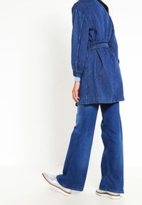 Jeansowa kurtka z paskiem w talii, długimi rękawami i luźnym krojem, w zestawie z szerokimi spodniami denimowymi. Oba w ciemnoniebieskim kolorze. Srebrne buty.