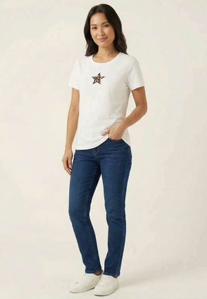 Donna in piedi che indossa una maglietta bianca con una stella stampata leopardata e jeans blu, scarpe da ginnastica bianche, con una mano in tasca.