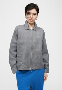 Νεαρός με κοντά σκούρα μαλλιά και γυαλιά που φοράει γκρι καρό μπουφάν με φερμουάρ και φωτεινό μπλε παντελόνι, σε απλό λευκό φόντο.