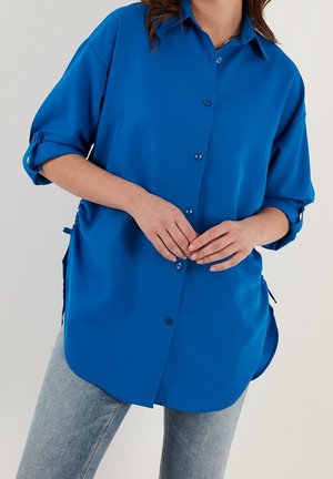 Kvinde iført en lys blå skjorte med knapper og opkrogede ærmer samt lyseblå jeans, stående med hænderne forsigtigt foldet.