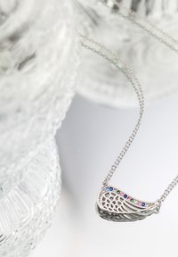 Sølv halskæde med en vingeformet vedhæng, prydet med farvede ædelsten og et detaljeret udskæringsdesign, præsenteret på en reflekterende overflade.