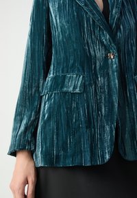 Teal velvet blazer featuring vertical stripes, a notched collar, a single button closure, and two front pockets. Soft, textured fabric.
