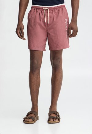 Uomo che indossa pantaloncini rosa con coulisse e scritta "Polo Club" sul lato e sandali marroni a doppio cinturino, in piedi davanti a uno sfondo neutro.