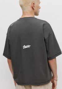 Oversized mørkegrå bomulds t-shirt med rund hals og et hvidt broderet logo på ryggen. Blød tekstur og afslappet pasform.