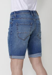 Denim šorti vidēji zilā krāsā, ar rūtiņotu apakšmalu, piecām kabatām, kontrastējošām diega šuvēm un ādas uzlīmi aizmugurējā jostā.