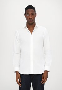 Camisa branca de manga longa feita de linho, com colarinho abotoado, botões frontais e uma textura suave. Combinada com jeans escuros.