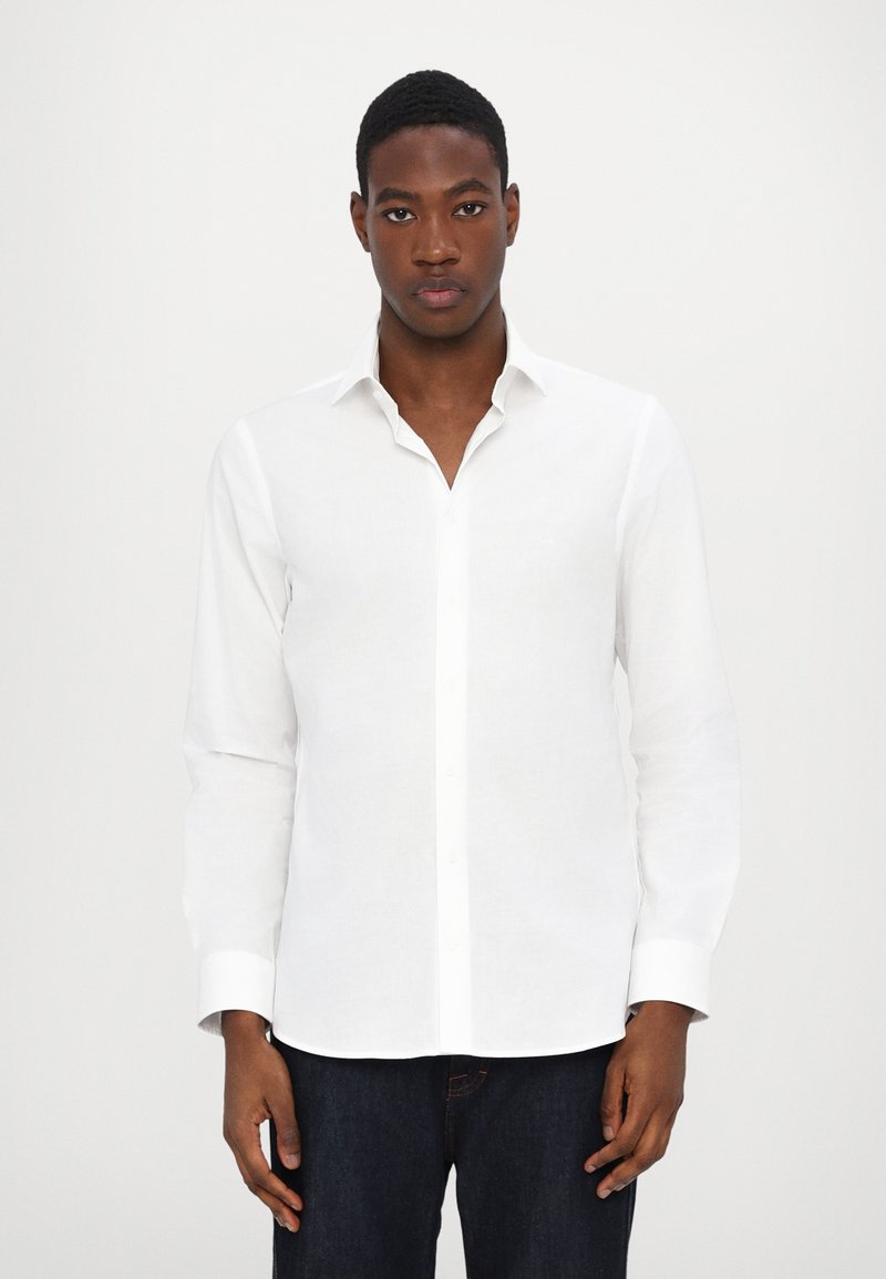 Camisa branca de manga longa feita de linho, com colarinho abotoado, botões frontais e uma textura suave. Combinada com jeans escuros.