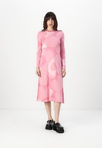 Rochie tricotată roz, cu modele abstracte de roz deschis, mâneci lungi, guler rotund și fustă de lungime medie. Asortată cu pantofi cu platformă negri.