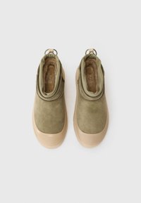 Olivgröna mocka ankelstövlar med en beige gummisula, som har en mjuk inre fodring, dragflik och en slät, rundad design.