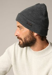 Bonnet tricoté gris avec bord côtelé et petite étiquette en cuir marron. Texture douce, coupe ajustée, forme effilée vers le haut.