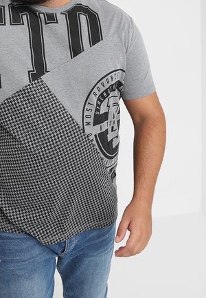 Homme portant un t-shirt gris à motifs noirs et un jean bleu, debout devant un fond blanc uni.