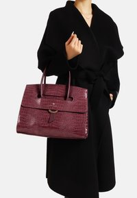 Borsa in tessuto coccodrillo color borgogna con doppio manico superiore, chiusura a pattina e un accento circolare, abbinata a un cappotto nero.