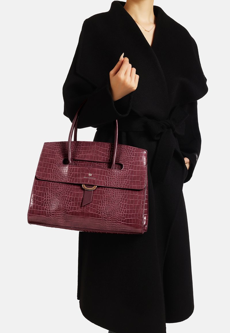 Borsa in tessuto coccodrillo color borgogna con doppio manico superiore, chiusura a pattina e un accento circolare, abbinata a un cappotto nero.