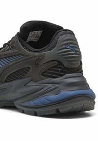 Melna un pelēka sporta kurpe ar strukturētu virsu, ziliem akcentiem, rakstainiem pārklājumiem un biezu zoli. Uz papēža ir redzams zīmola logo.