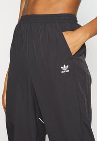 Personne portant un pantalon de survêtement noir Adidas avec une ceinture élastique, main dans la poche droite, et logo Adidas sur la cuisse supérieure.