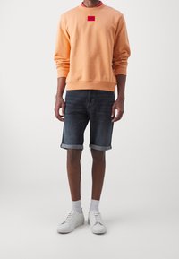 Pomarańczowa bluza z kapturem z czerwoną naszywką z logo, zestawiona z ciemnymi szortami dżinsowymi z podwiniętymi mankietami oraz białymi trampkami. Prosty design, casualowy krój.