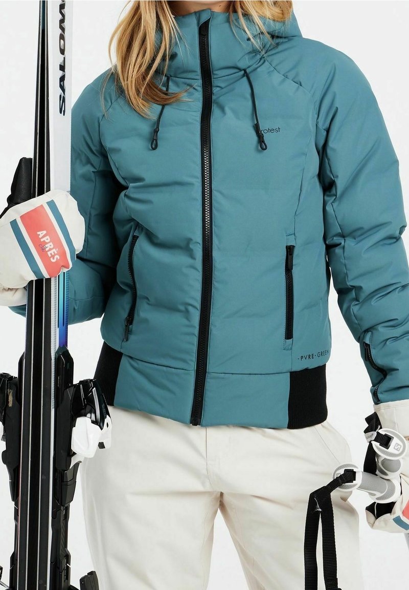 Türkisfarbene isolierte Jacke mit Reißverschluss vorne, schwarzen Akzenten und verstellbarem Kapuze. Das Model hält Skier und trägt weiße und blaue Handschuhe.