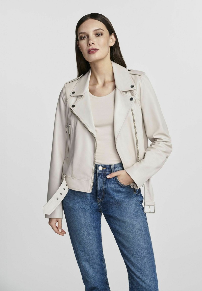 Jasny, dopasowany skórzany kurtka z zamykanymi kieszeniami i guzikami, noszona na beżowej bluzce i zestawiona z niebieskimi dżinsami.