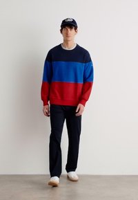 Color-blocked sweater featuring blue, red, and navy sections. Paired with dark trousers and white sneakers. Casual cap completes the look.