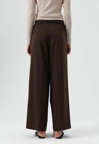 Bruine broek met wijde pijpen en een gladde textuur, voorzien van een hoge taille, twee achterzakken en een getailleerde pasvorm. Gecombineerd met een beige geribbelde top.