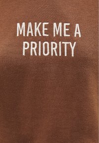 Tela marrón con texto bordado en blanco que dice "CONVIÉRTEME EN UNA PRIORIDAD" centrado en letras mayúsculas.