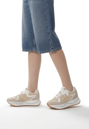 Ben iført blå cropped denimbukser og beige-hvide sneakers med teksturerede sider og tykke hvide såler, stående på en hvid overflade.