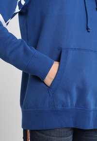 Blå hoodie med dragsnöre, framficka och långa ärmar. Materialet verkar mjukt med en slät textur och kontraststickning.