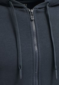 Mörkgrå zip-up hoodie i strukturerad tyg, med en framzipper med branding, justerbara dragsnören och en minimalistisk design.