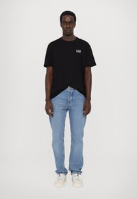 Svart bomull t-skjorte med en liten hvit "EA7" logo på venstre bryst, kombinert med lyseblå denimjeans og hvite sneakers.