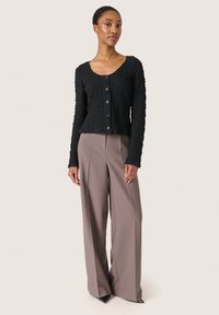 Cardigan nero con texture, maniche lunghe e bottoni frontali, abbinato a pantaloni taupe a vita alta e gamba larga, e scarpe nere a punta.