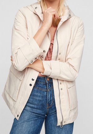 Žena v světle béžové bundě s kapucí přes růžový krajkový top a modré džínové kalhoty, s překříženýma rukama a rukou u brady.