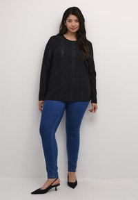 Schwarzes Langarmshirt mit gewelltem Cut-out-Design, kombiniert mit blauen Skinny-Jeans und schwarzen Slingback-Heels mit spitzem Zehen.