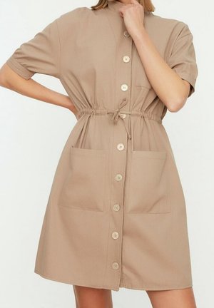 Beige mekko, joka on valmistettu pehmeästä kankaasta, lyhyillä hihoilla, nappi etuosassa, kahdella etutaskulla ja säädettävä naru vyötäröllä.