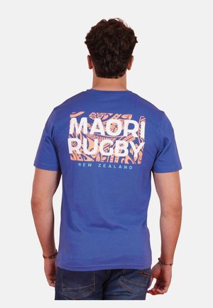 Homme portant un T-shirt bleu avec un motif maori orange et le texte blanc "Maori Rugby Nouvelle-Zélande" au dos, tourné de dos.
