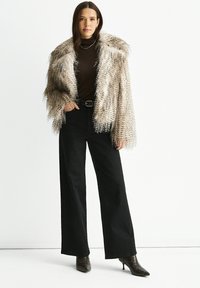 Woman wearing a beige faux fur jacket, black turtleneck, black wide-leg pants, black belt, and pointed-toe heeled boots standing against white background.