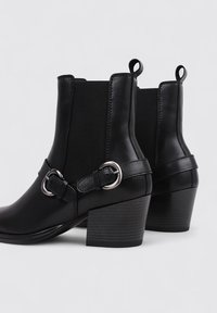 Bottines noires en cuir lisse avec une finition matte. Elles présentent un talon rectangulaire, des accents en métal argenté et des panneaux élastiques sur les côtés.