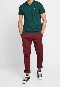 Mężczyzna w zielonej koszulce polo, burgundowych spodniach z podwiniętymi nogawkami i czarnych niskich trampkach, stojący z rękami w kieszeniach na białym tle.