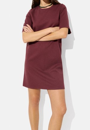 Woman wearing a short-sleeve, burgundy, knee-length dress with a round neckline and green braided necklace, arms crossed.