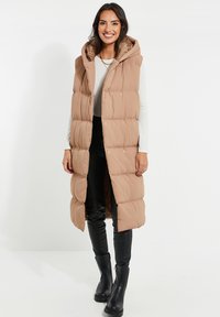 Long gilet isolé de couleur brun clair avec une capuche. Présente un design matelassé et des accents en métal doré. Porté sur un pantalon noir et des bottes.