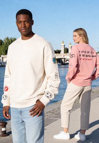 Jeune homme en sweat-shirt blanc à motif graphique et jean près du bord de l'eau, femme en sweat-shirt rose avec texte pose de dos.